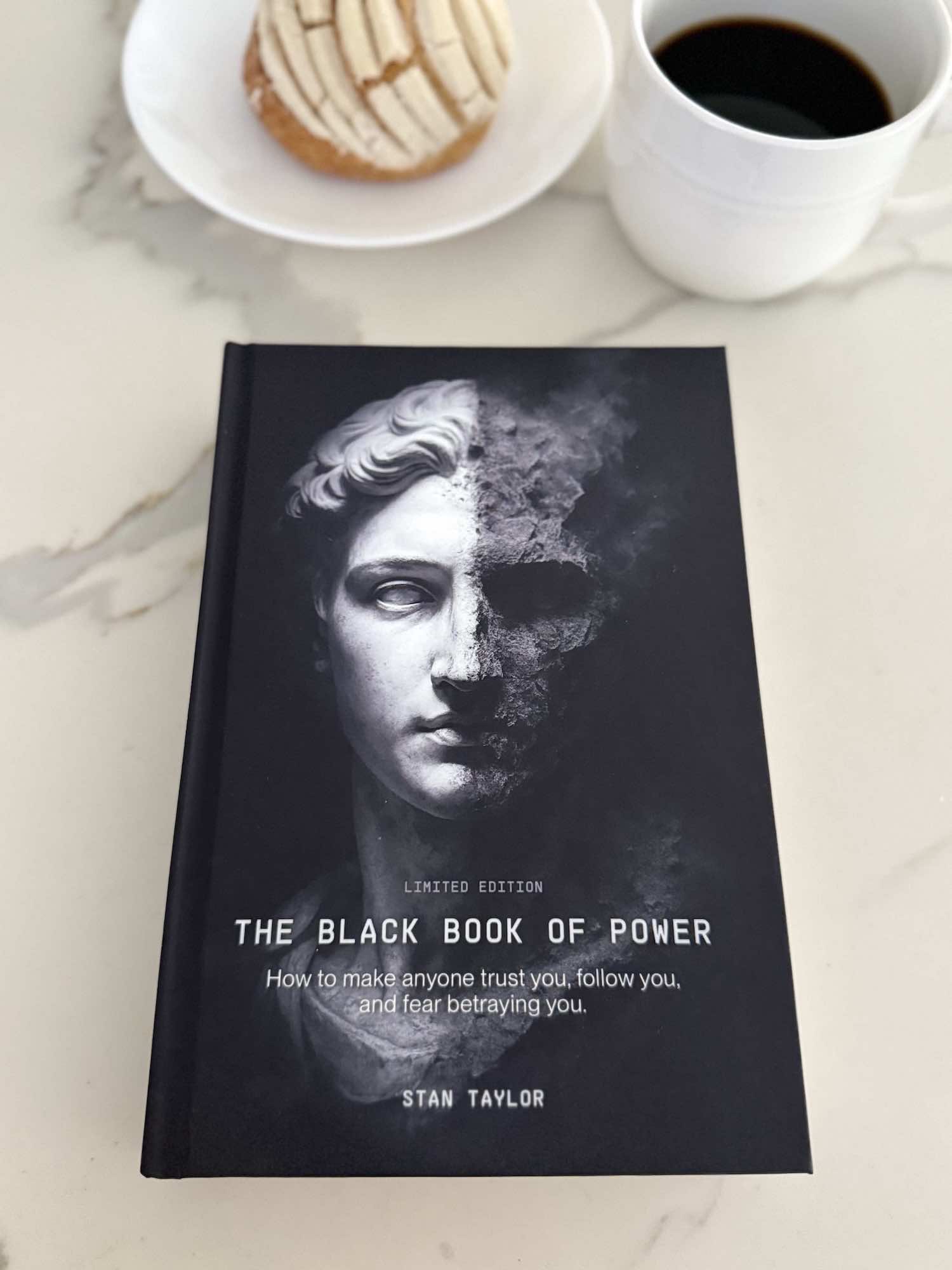 Book titled 'The Black Book of Power' on a marble surface with a cup of coffee and cookies.