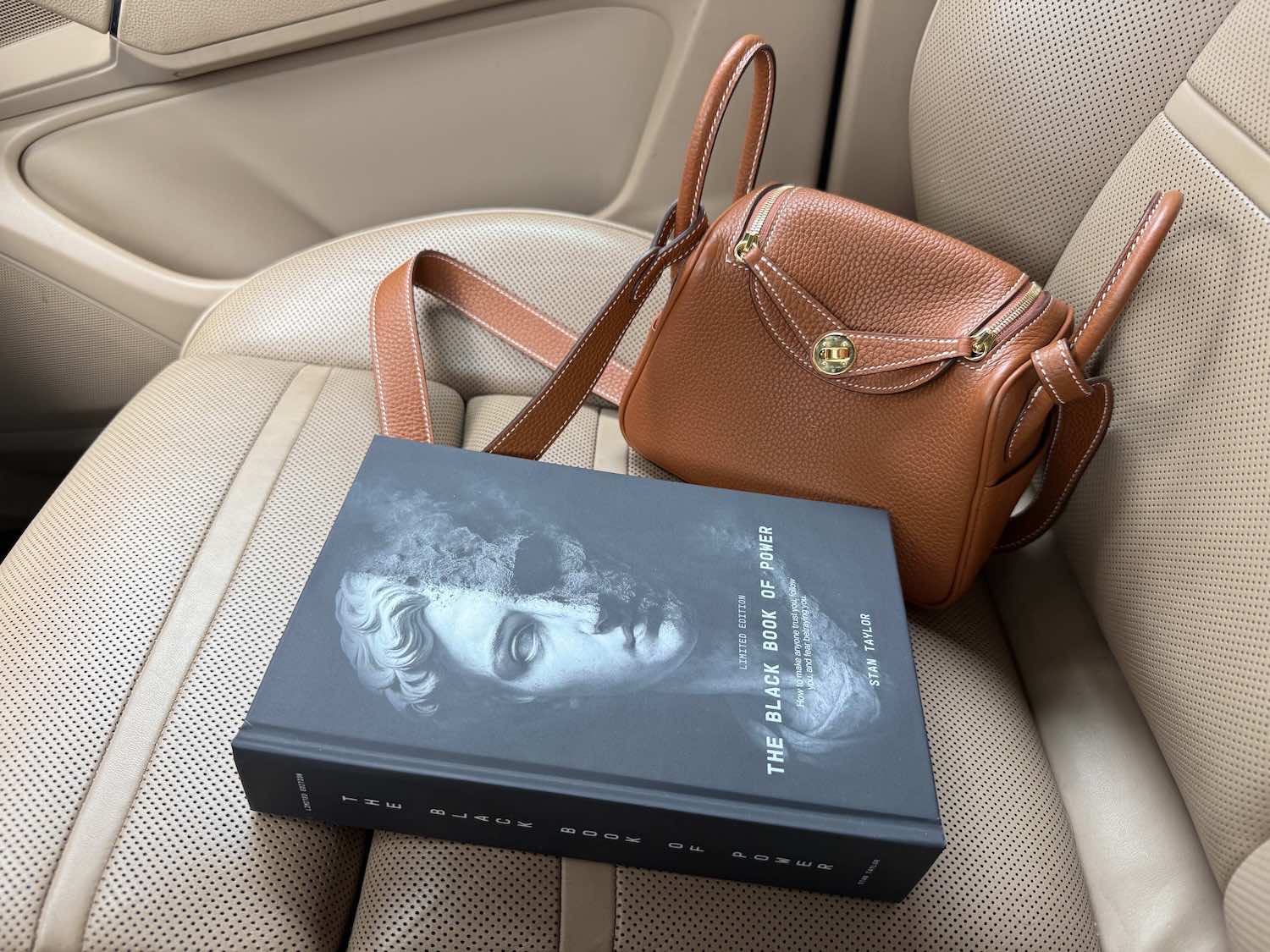 Brown leather handbag and book titled 'The Black Book of Power' on a car seat.